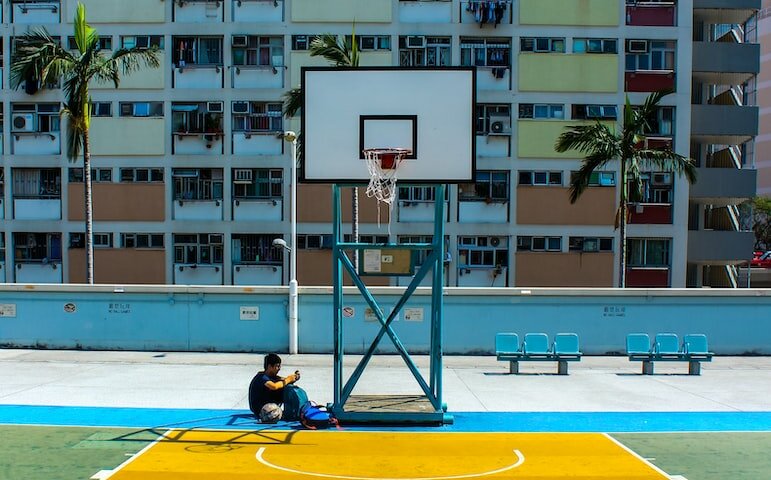 man sitting below basketball hoop near the building during daytime